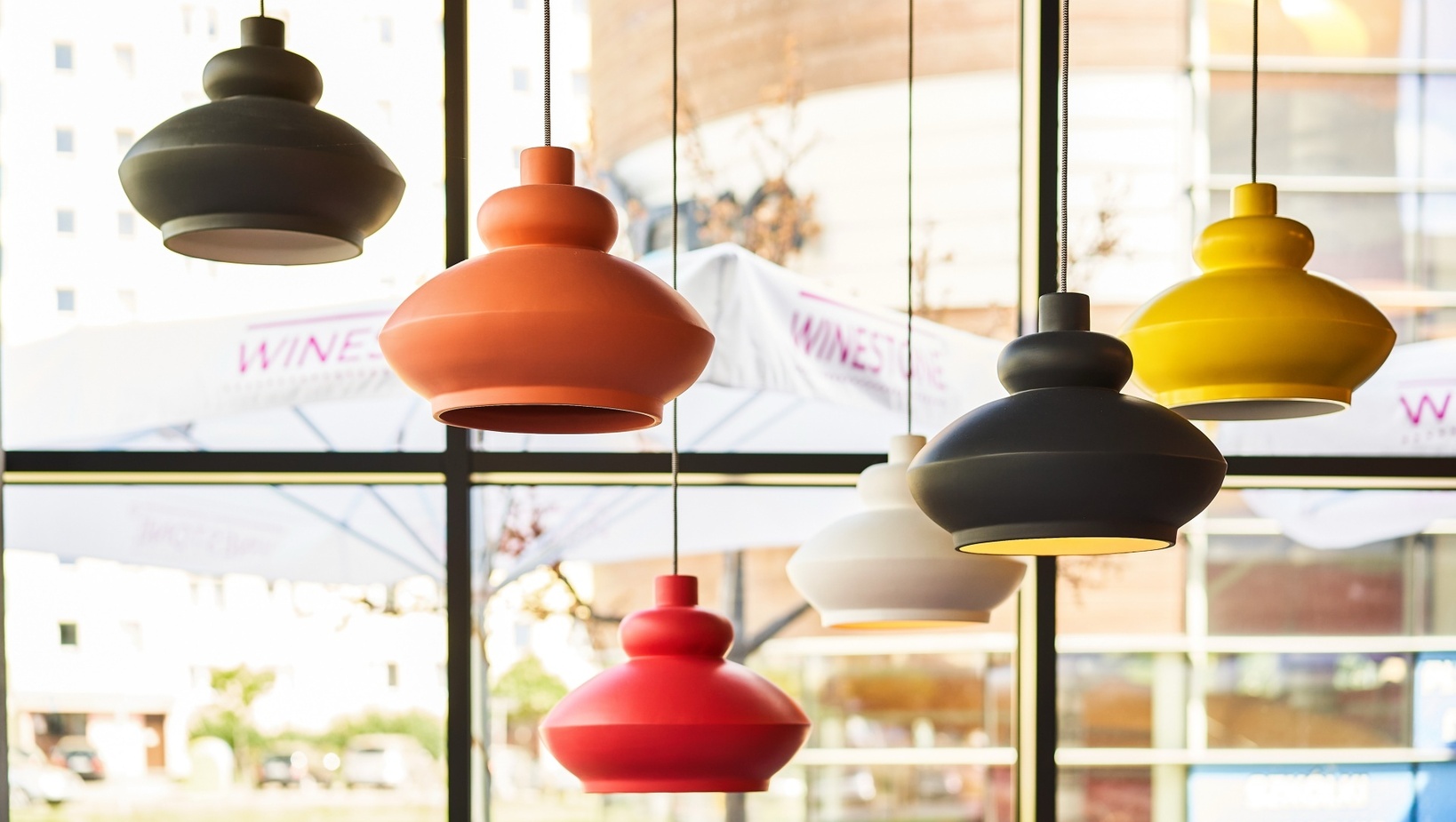 Kolorowe, nowoczesne lampy wiszące nad częścią restauracyjną; duże okna i zewnętrzne parasole na tarasie.
