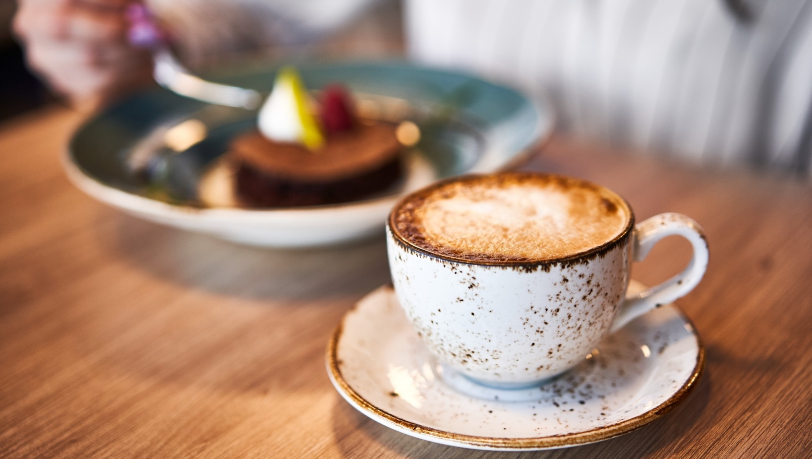 Kremowe cappuccino w cętkowanej filiżance na drewnianym stoliku, w tle rozmyte ciasto czekoladowe z owocami.