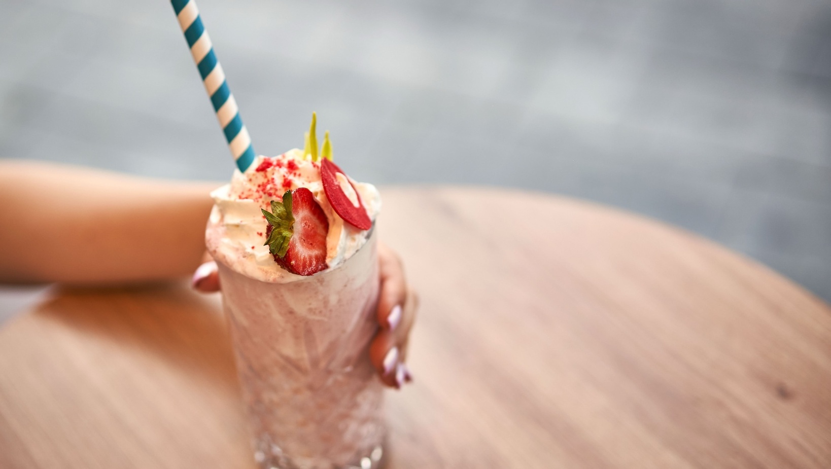 Kremowy milkshake truskawkowy z bitą śmietaną, plasterkiem truskawki i niebiesko-białą papierową słomką w dekoracyjnej szklance na drewnianym stoliku.
