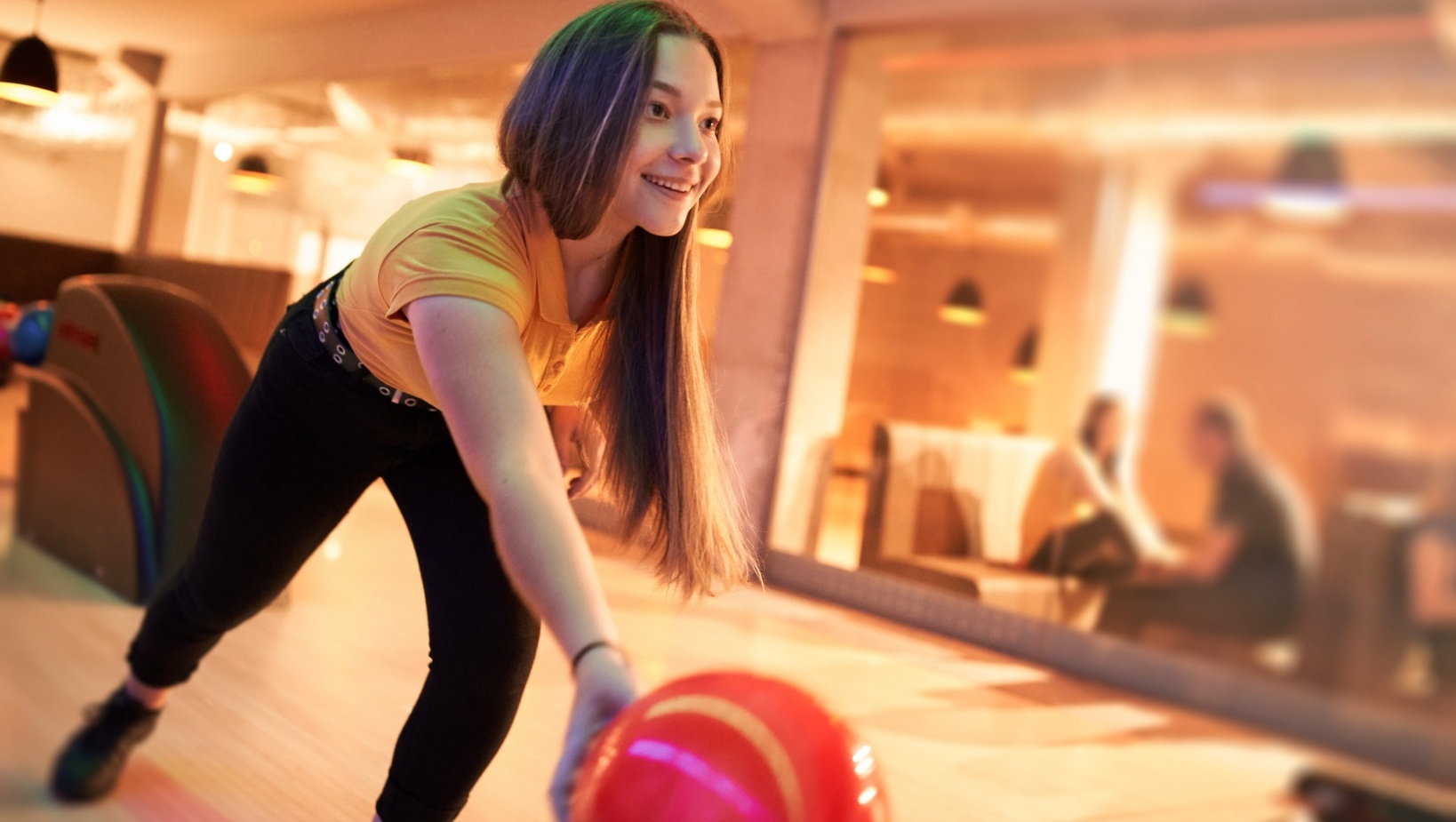 Młoda kobieta rzucająca czerwoną kulą na torze bowlingowym w hotelowej rozrywce, przytulne oświetlenie i strefa lounge w tle
