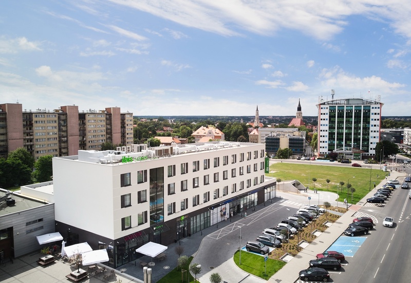 Widok na nowoczesny hotel Ibis Styles z tarasem kawiarnianym i parasolami, parkingiem przed hotelem oraz panoramą miasta z wieżami kościołów.