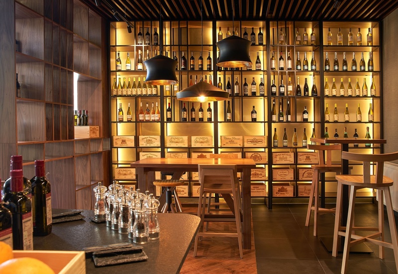 Elegancki bar i winiarnia hotelowa z drewnianymi regałami pełnymi butelek, podświetlonymi skrzyniami wina, stołem, hokerami i wiszącymi lampami.
