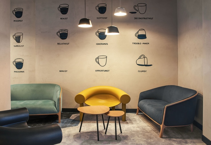 Przytulny lounge z zielonym, żółtym i granatowym fotelem, trzema stolikami, wiszącymi lampami i muralem filiżanek na ścianie.