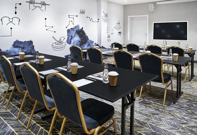 Sala konferencyjna z stołami i krzesłami, dużym monitorem, butelkami wody i kubkami z kawą, nowoczesna grafika na ścianie.
