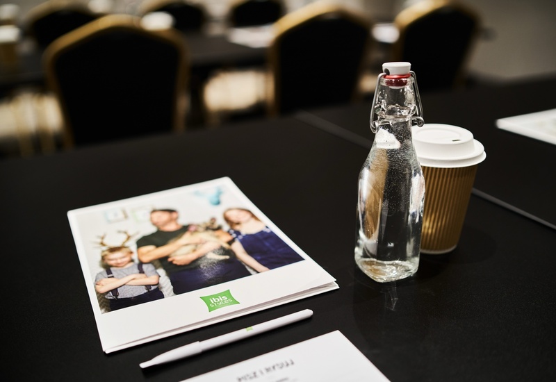 Stół konferencyjny z butelką wody, papierowym kubkiem kawy, notesem Ibis Styles i długopisem, rozmyte krzesła w tle