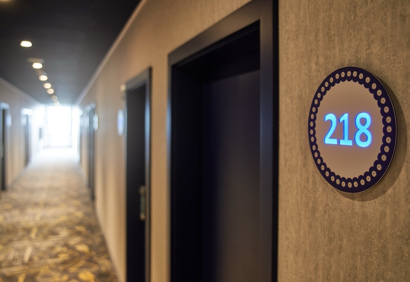 Korytarz hotelowy z miękkim oświetleniem sufitowym, wzorzystą wykładziną i podświetloną tabliczką pokoju 218 przy nowoczesnych drzwiach.
