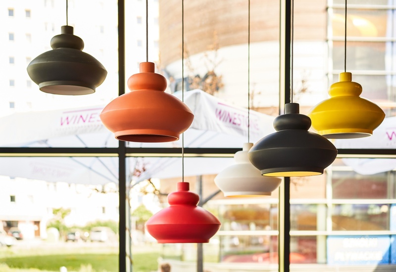Kolorowe, nowoczesne lampy wiszące nad częścią restauracyjną; duże okna i zewnętrzne parasole na tarasie.