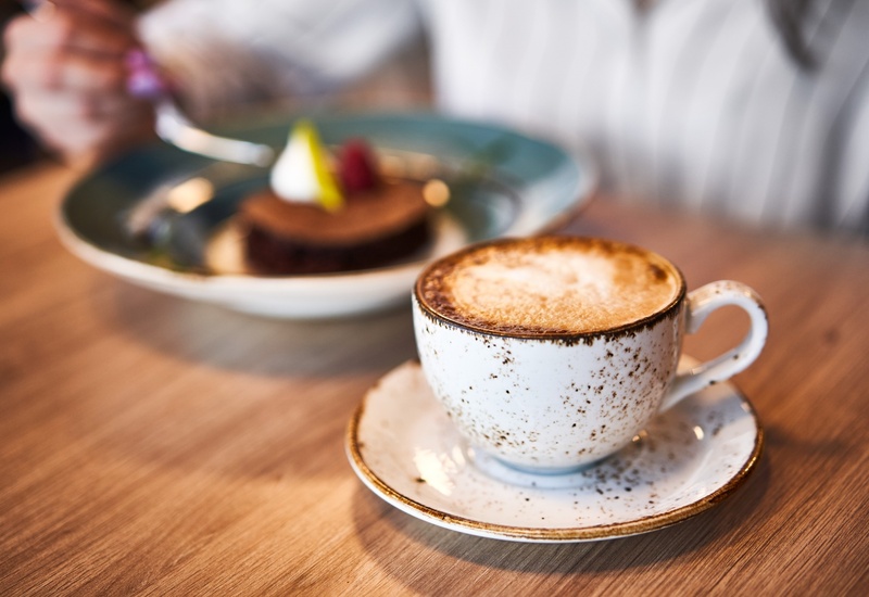Kremowe cappuccino w cętkowanej filiżance na drewnianym stoliku, w tle rozmyte ciasto czekoladowe z owocami.