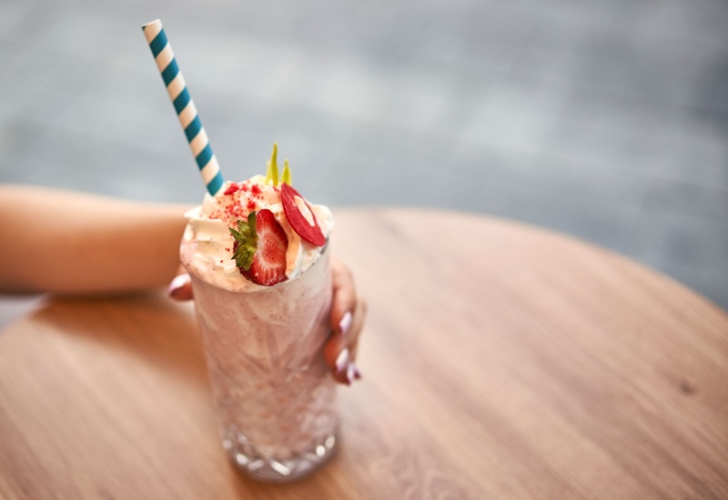Kremowy milkshake truskawkowy z bitą śmietaną, plasterkiem truskawki i niebiesko-białą papierową słomką w dekoracyjnej szklance na drewnianym stoliku.
