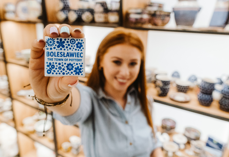 Ręka z pastelowym manicure trzyma ceramiczną pamiątkę „Bolesławiec” na tle półek z dekoracyjną ceramiką w sklepie.