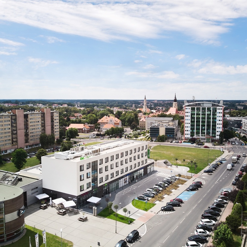 Nowoczesny hotel Ibis Styles z tarasem i stolikami na zewnątrz, parkingiem; w tle miasto z wieżami kościołów i zielonymi parkami