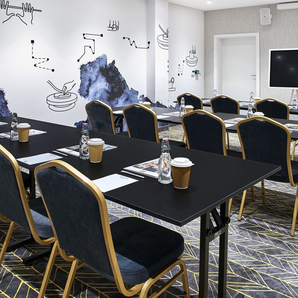 Sala konferencyjna hotelu w układzie szkoleniowym z czarnymi stołami, granatowymi krzesłami, ekranem, wodą i kawą.