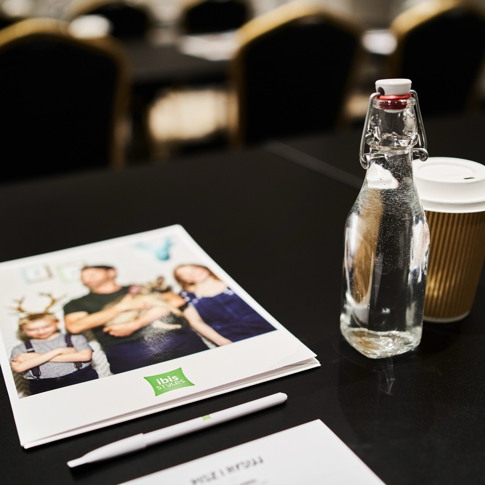 Stół konferencyjny z butelką wody, papierowym kubkiem kawy, notesem Ibis Styles i długopisem, rozmyte krzesła w tle