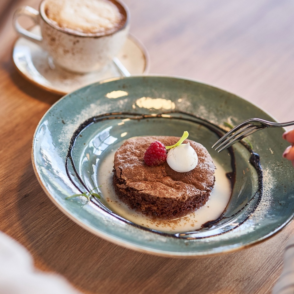 Czekoladowe ciastko na ceramicznym talerzu z kremową bezą, maliną i sosem, obok filiżanka cappuccino i dłoń z widelcem.