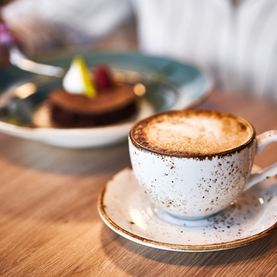 Kremowe cappuccino w cętkowanej filiżance na drewnianym stoliku, w tle rozmyte ciasto czekoladowe z owocami.