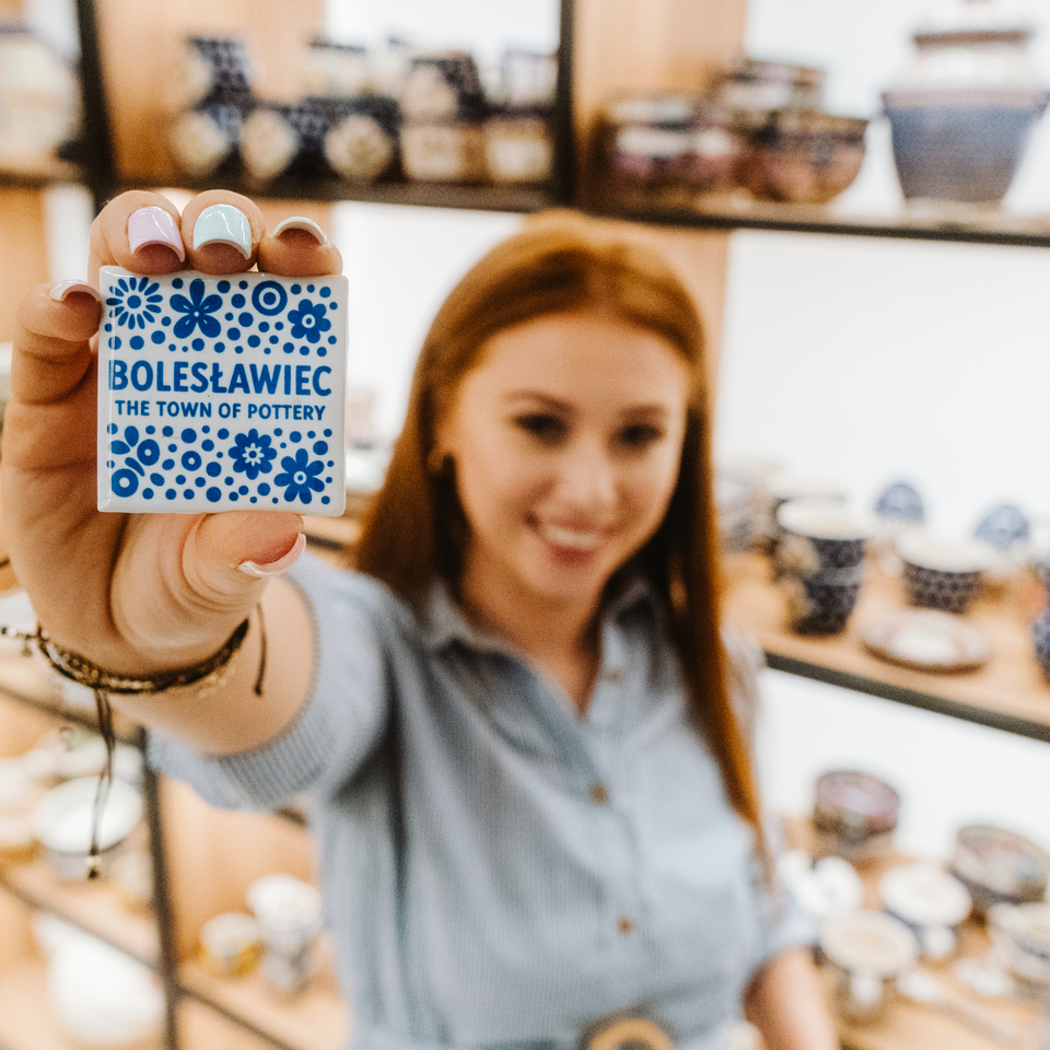 Ręka z pastelowym manicure trzyma ceramiczną pamiątkę „Bolesławiec” na tle półek z dekoracyjną ceramiką w sklepie.