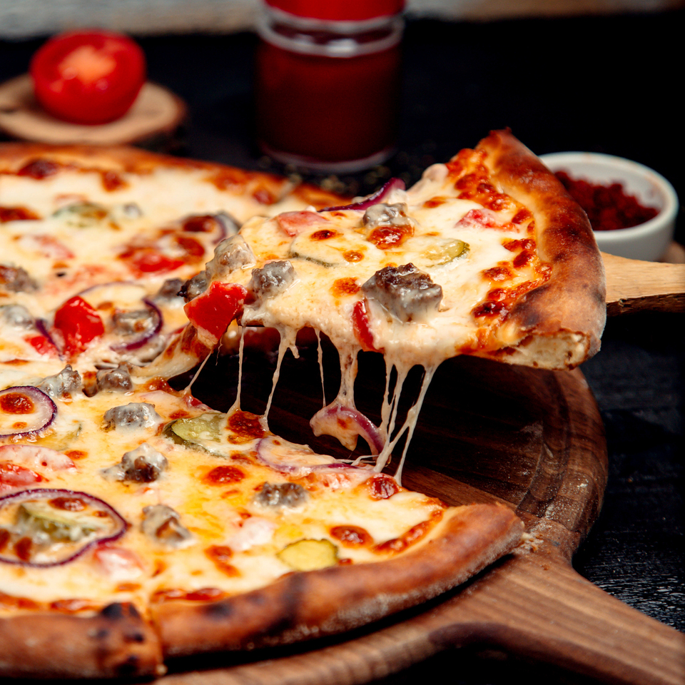 Kawałek pizzy z ciągnącym się serem, kiełbasą, pomidorkami i cebulą na złocistej skórce, podany na drewnianej desce