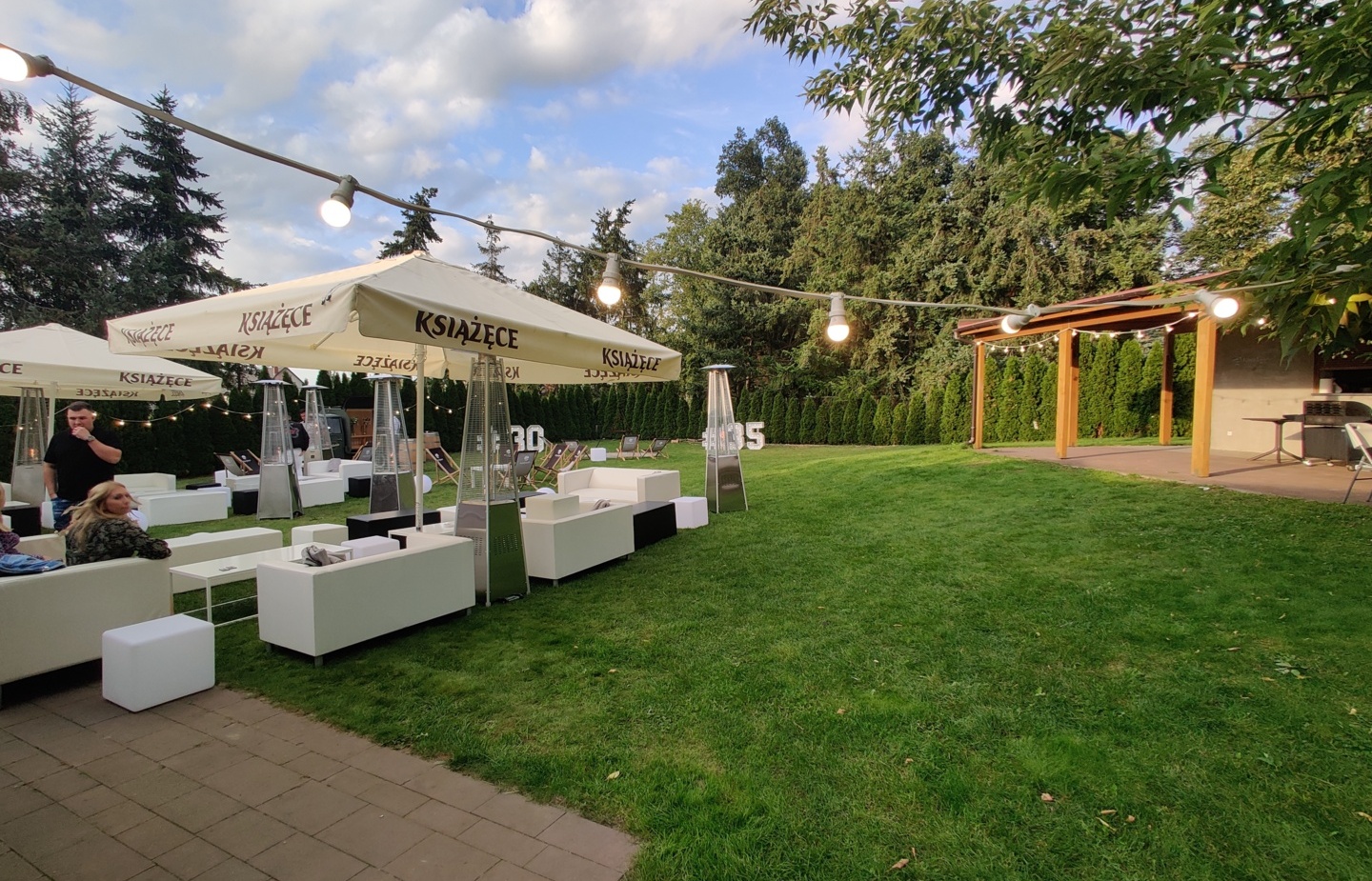 Zewnętrzny salon na trawniku z białymi sofami pod parasolami Książęce, lampkami, nagrzewnicami i drewnianą pergolą.