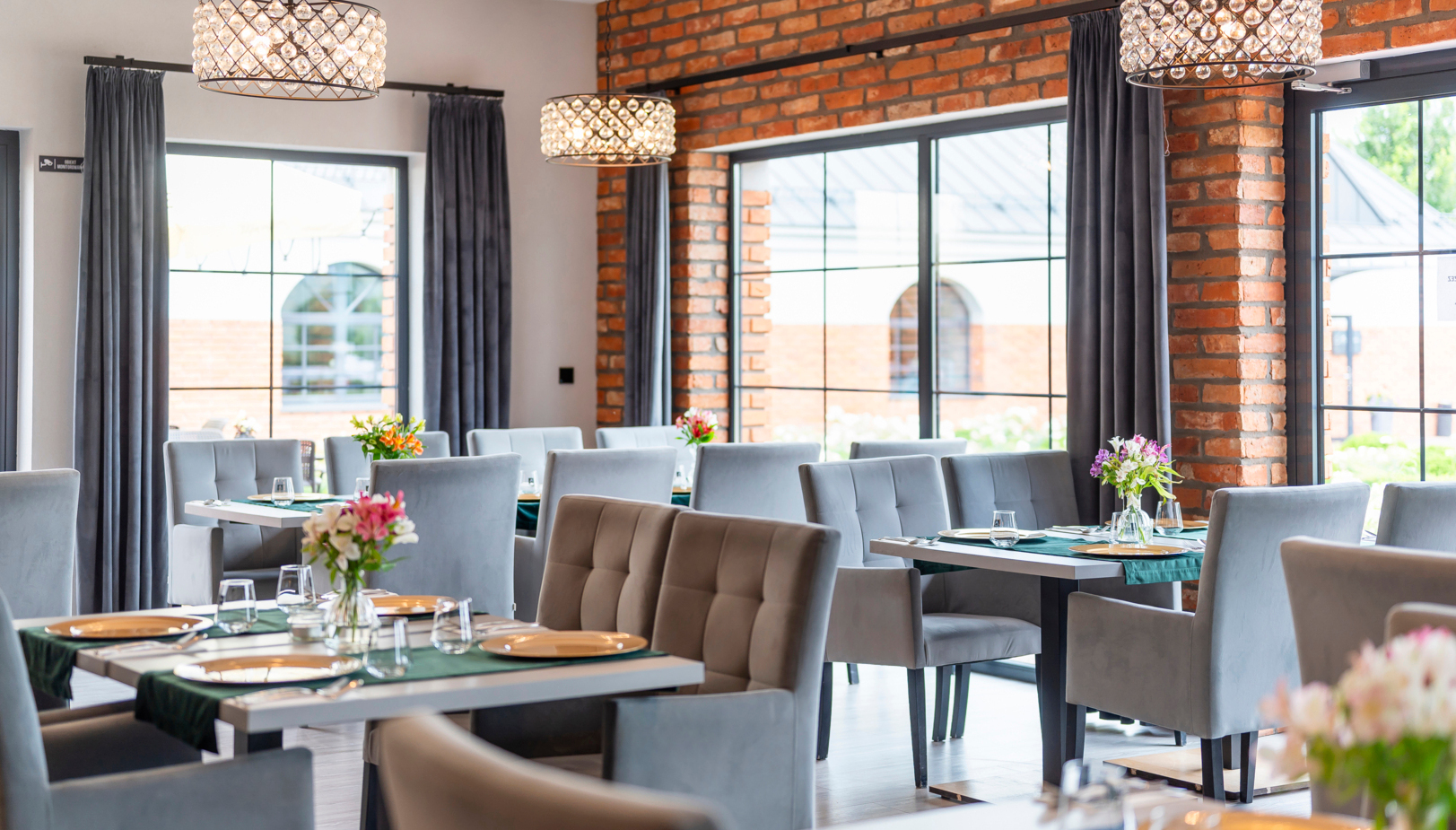 Jasna restauracja hotelowa z ceglaną ścianą, dużymi oknami, kryształowymi lampami wiszącymi, szarymi welwetowymi fotelami i kwiatami na stołach
