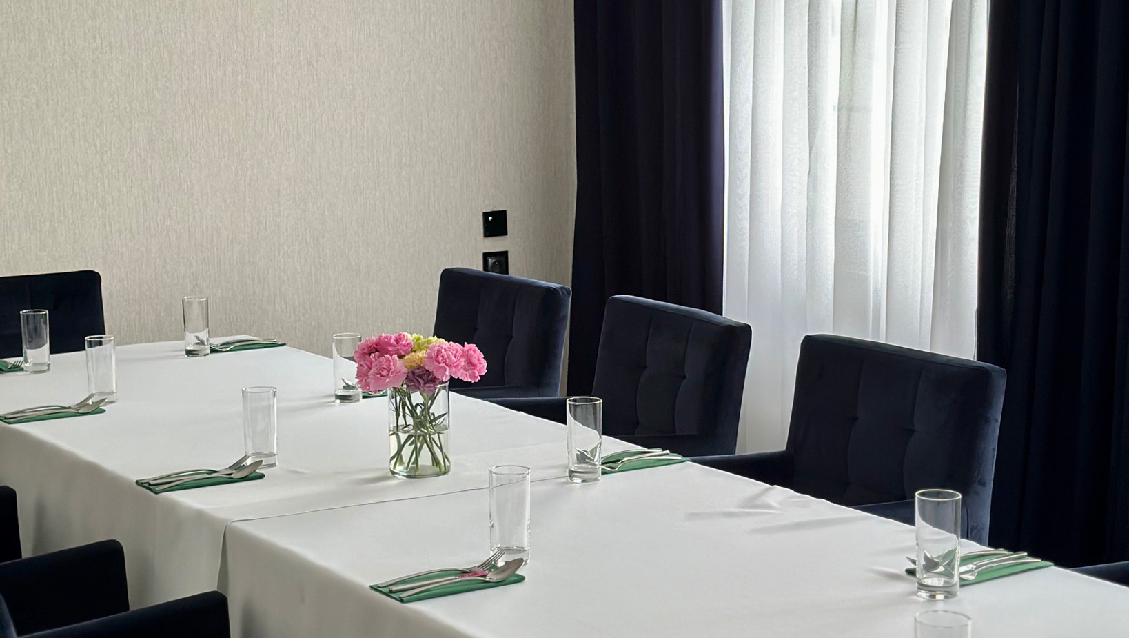 Sala konferencyjna z długim stołem w białym obrusie, granatowymi fotelami, wazonem różowych kwiatów i zwiewnymi firankami.