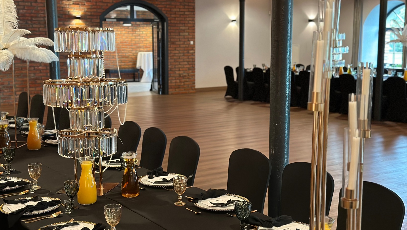 Elegancka sala bankietowa z czarnymi obrusami, kryształową piętrową dekoracją stołu, karafkami soku pomarańczowego, ceglanym murem i łukowym przejściem.