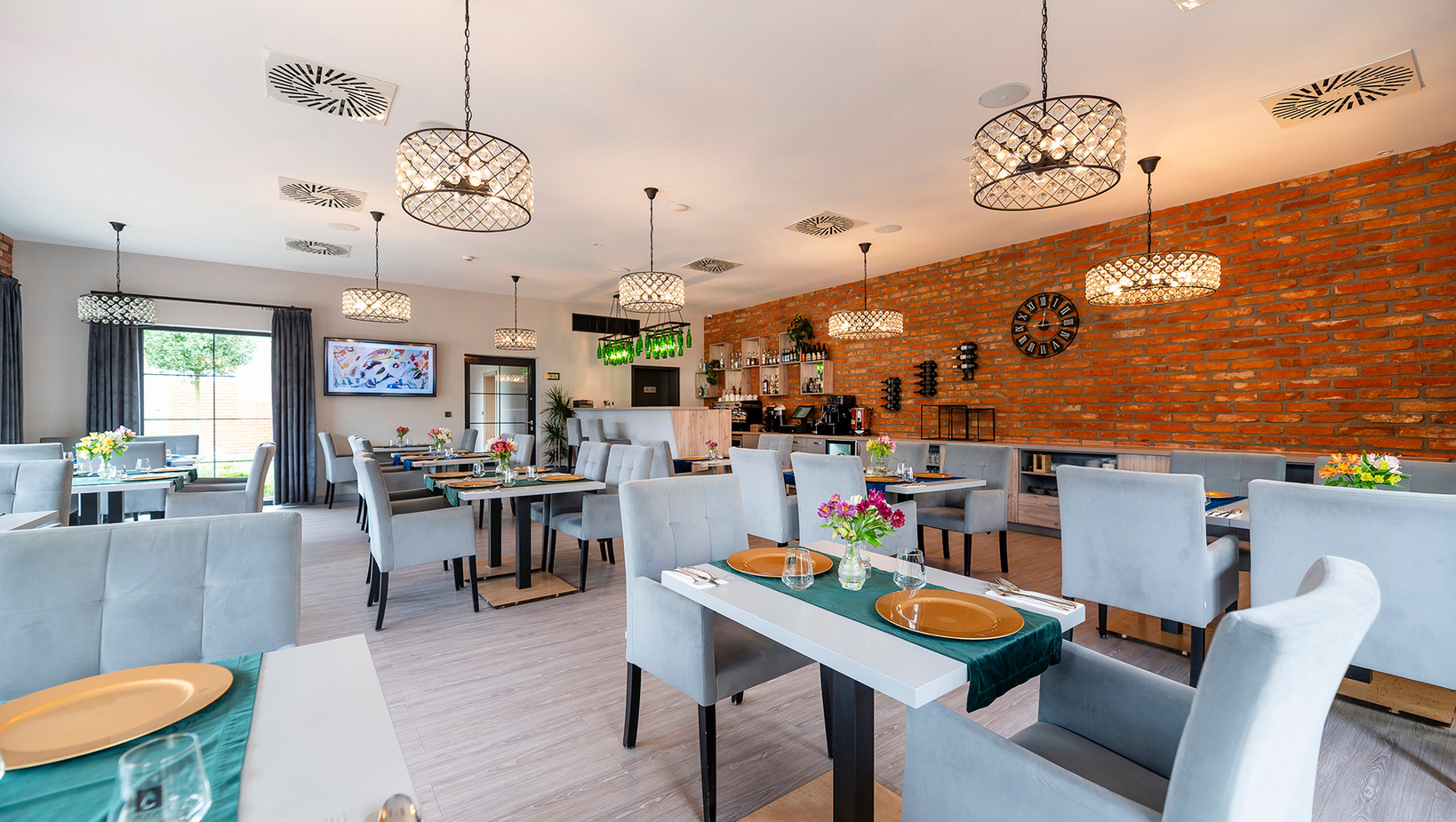 Nowoczesna restauracja hotelowa z szarymi fotelami, białymi stołami, turkusowymi akcentami, kryształowymi lampami i ceglaną ścianą.