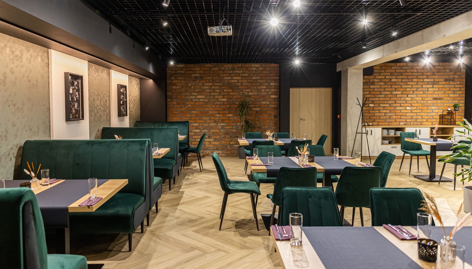 Restauracja hotelowa z zielonymi aksamitnymi kanapami, nakrytymi drewnianymi stołami, ceglanym murem i ciepłym oświetleniem