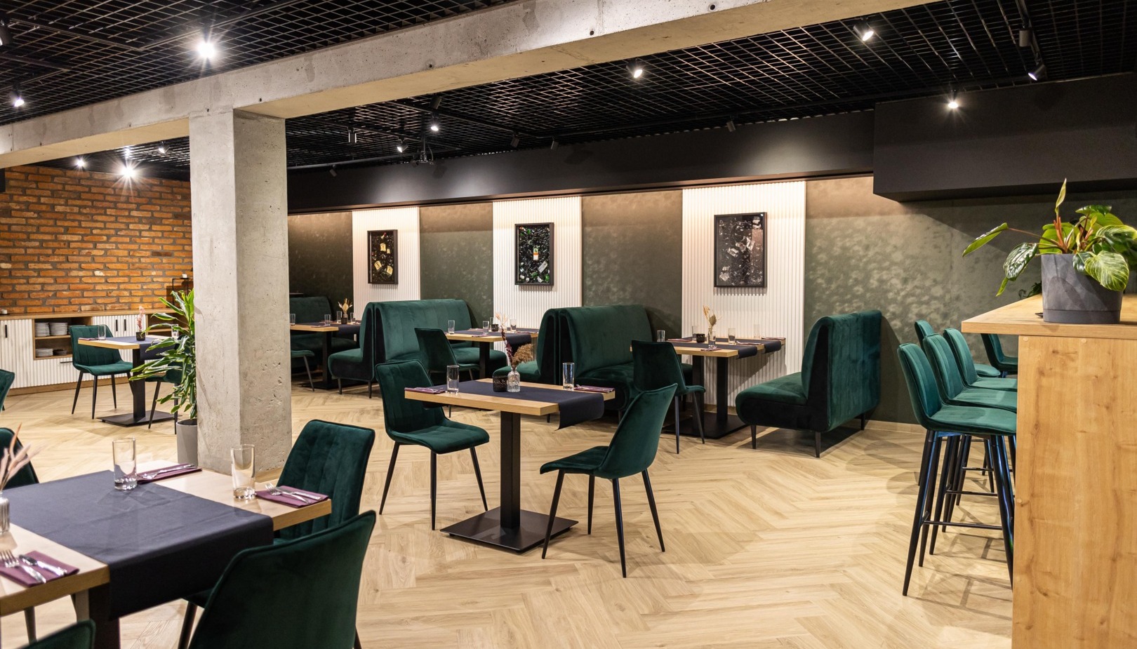 Restauracja hotelowa z zielonymi aksamitnymi boksami i krzesłami, podłogą w jodełkę, nakrytymi stolikami, ceglaną ścianą i ciepłym światłem