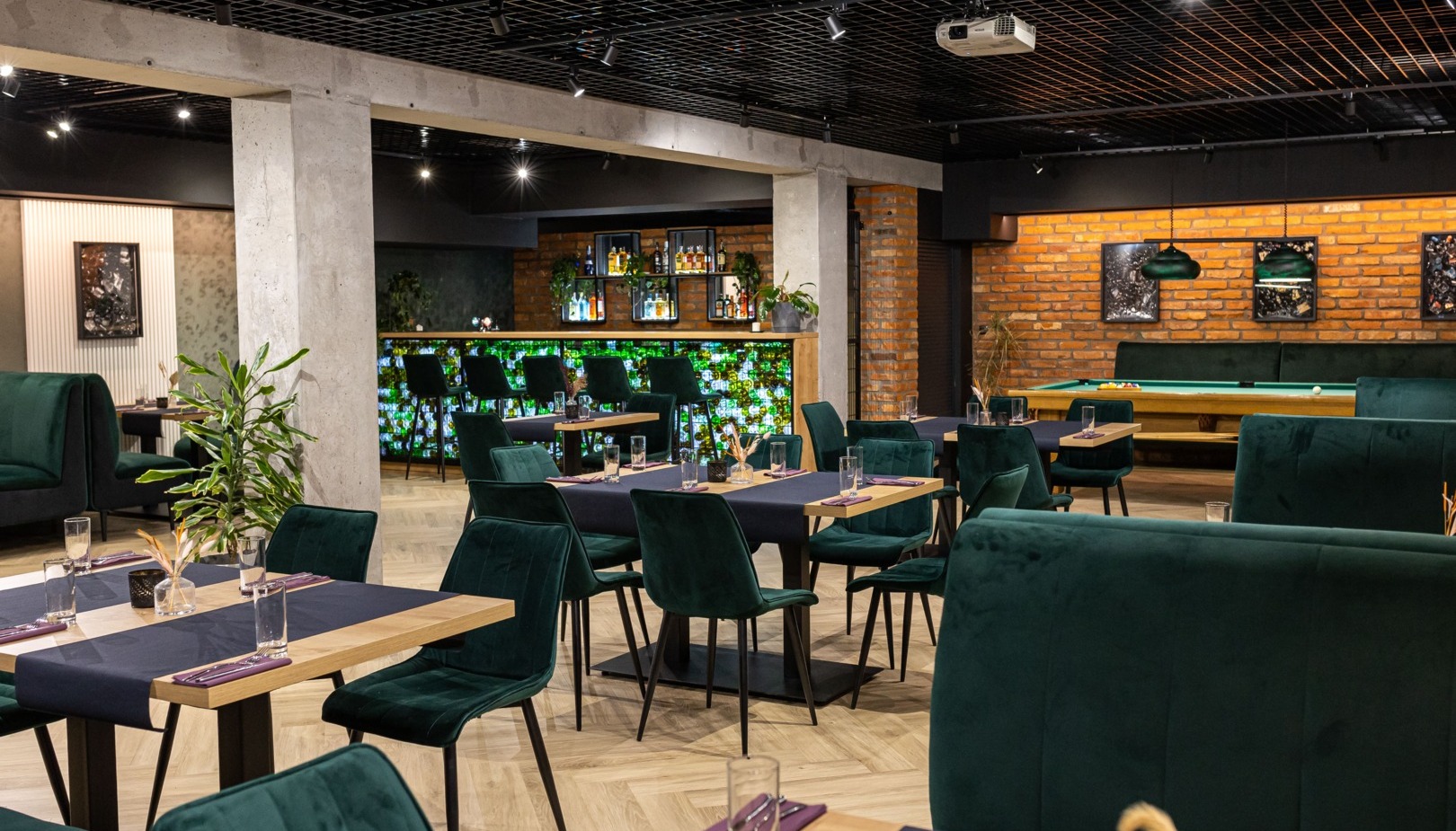 Stylowy lounge hotelowy z zielonymi aksamitnymi siedziskami, drewnianymi stolikami, podświetlonym barem i stołem bilardowym przy ceglanej ścianie.