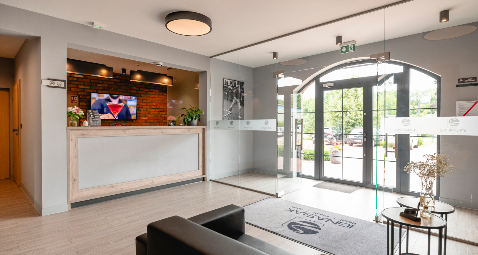 Bright, modern hotel lobby with reception in a wooden frame, exposed brick wall, glass arched entrance, sofas, and plants.