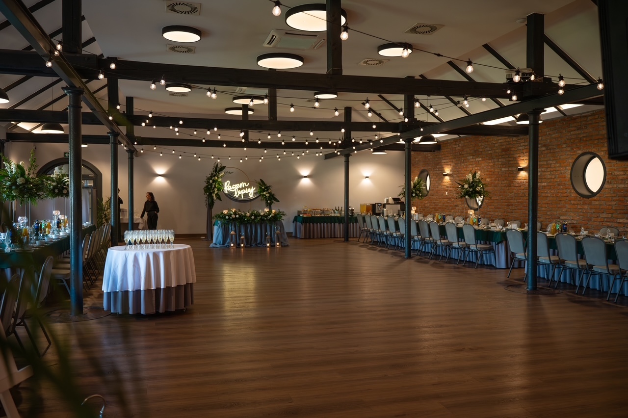 Spacious banquet hall with exposed beams, wooden floor, light garlands, long tables, buffet, and flowers.