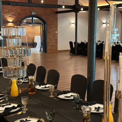Elegancka sala bankietowa z czarnymi obrusami, kryształową piętrową dekoracją stołu, karafkami soku pomarańczowego, ceglanym murem i łukowym przejściem.