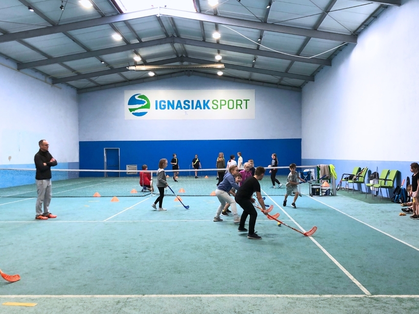 Wewnętrzna hala sportowa, dzieci grają w unihokeja pod opieką trenera na korcie z siatką, miejsca siedzące i sprzęt dla rodzinnych zajęć.