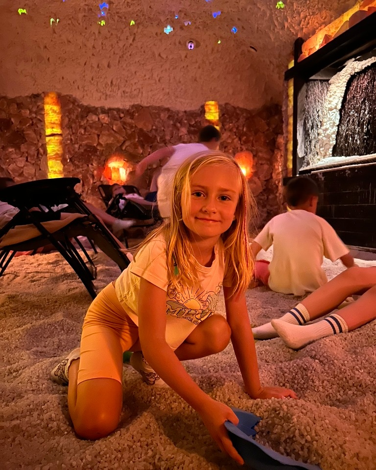 Salt-grotto relaxation room with warm amber lamps, starry ceiling and soft salt floor; children playing with scoops by a salt waterfall.