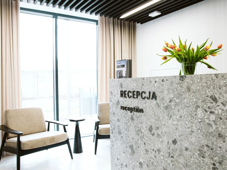 Hotel reception with mottled stone desk reading RECEPCJA/reception, tulips in glass vase, lounge chairs and large windows