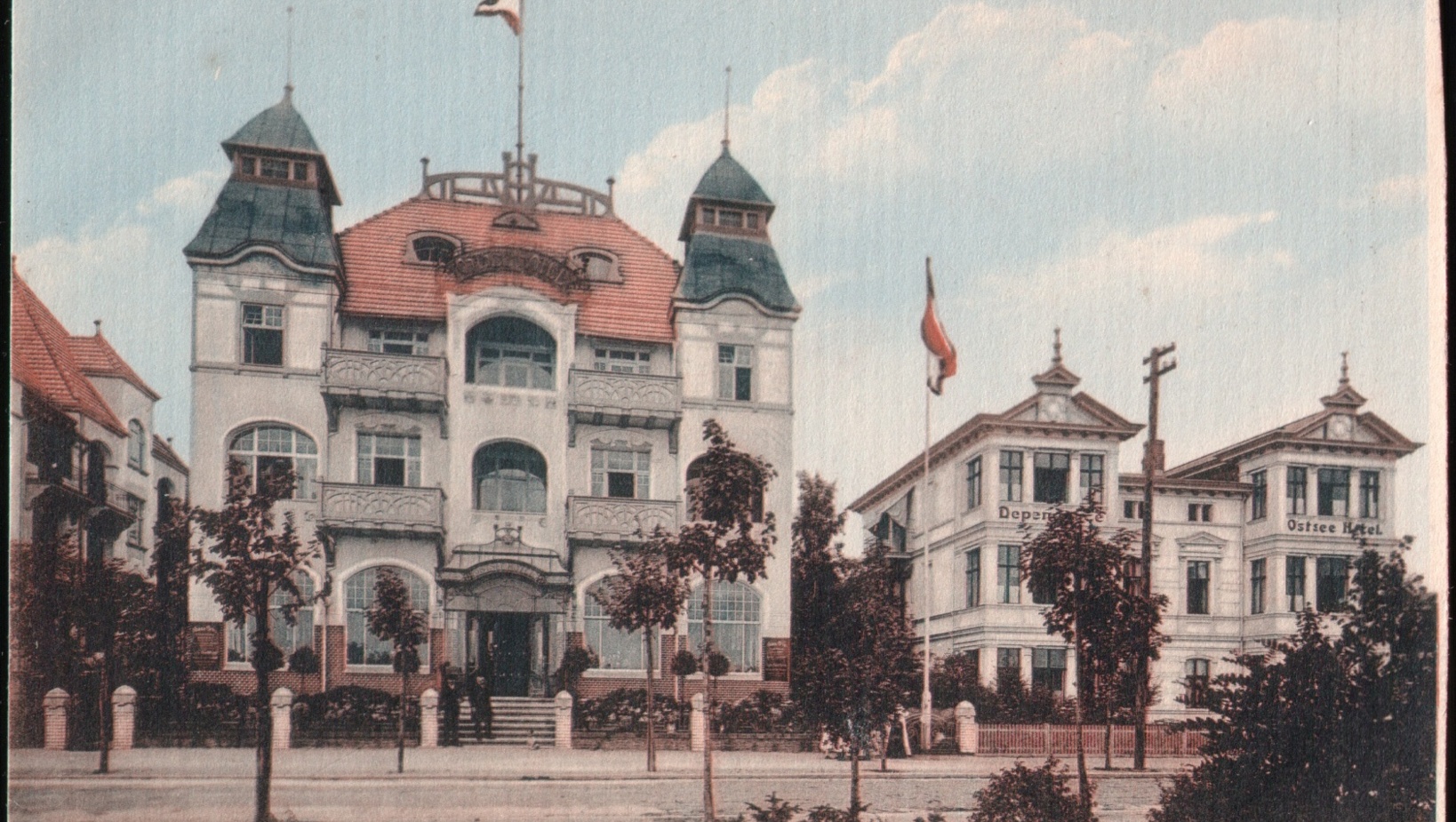 Trzywieżowy hotel o klasycznej architekturze z czerwonym dachem i zielonymi wieżyczkami, otoczony drzewami i ogrodzeniem.