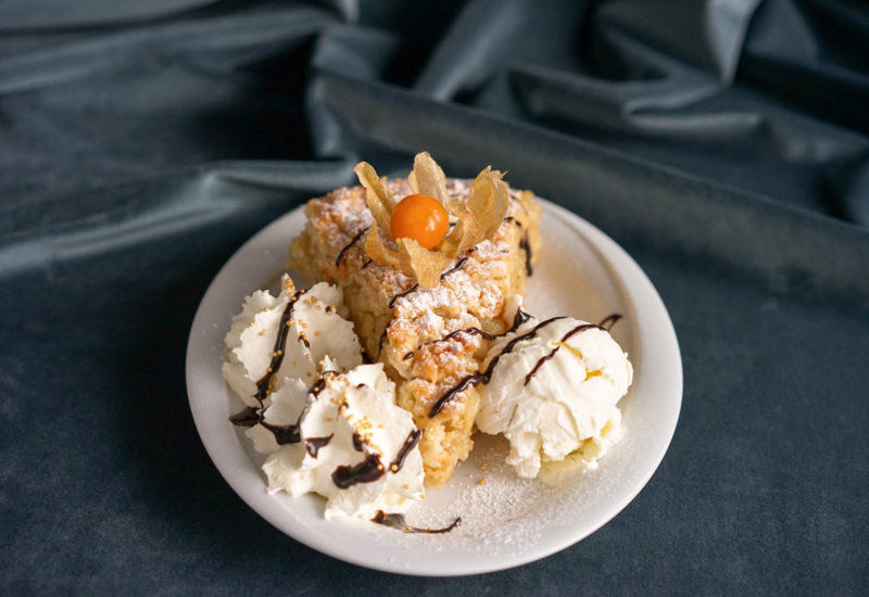 Porcja ciasta z owocem physalis, podana z bitą śmietaną i gałką lodów, polane czekoladą na białym talerzu.