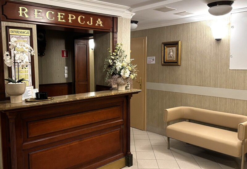 Drewniana recepcja Wellness z marmurowym blatem, storczykiem i dekoracją kwiatową, beżowa kanapa, ciepłe światło i tablica informacji.