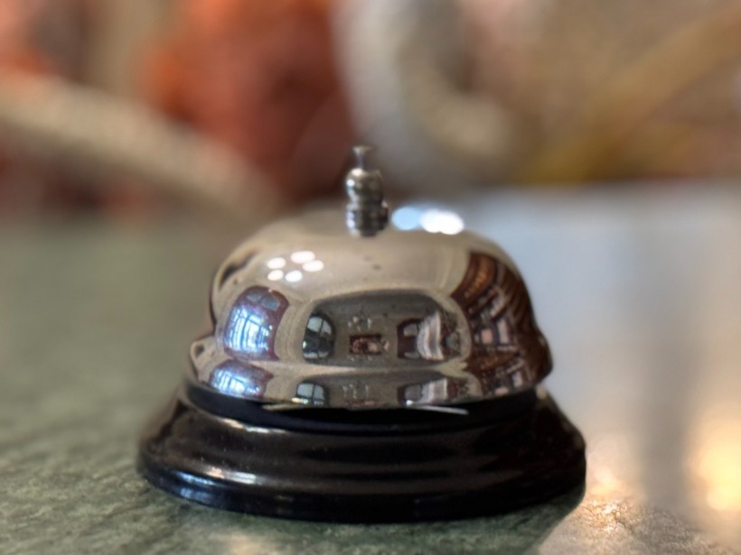 Metalowy dzwonek recepcyjny na blacie, odbijający jasne wnętrze hotelu w tle.