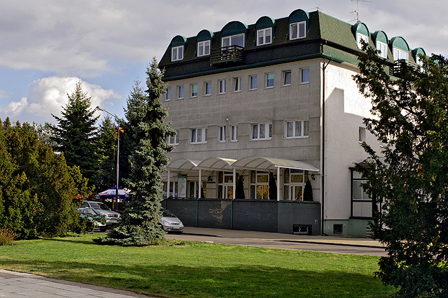 Trzypiętrowy hotel z zielonym dachem i oknami na poddaszu, otoczony drzewami i trawnikiem, z zadaszonym wejściem.