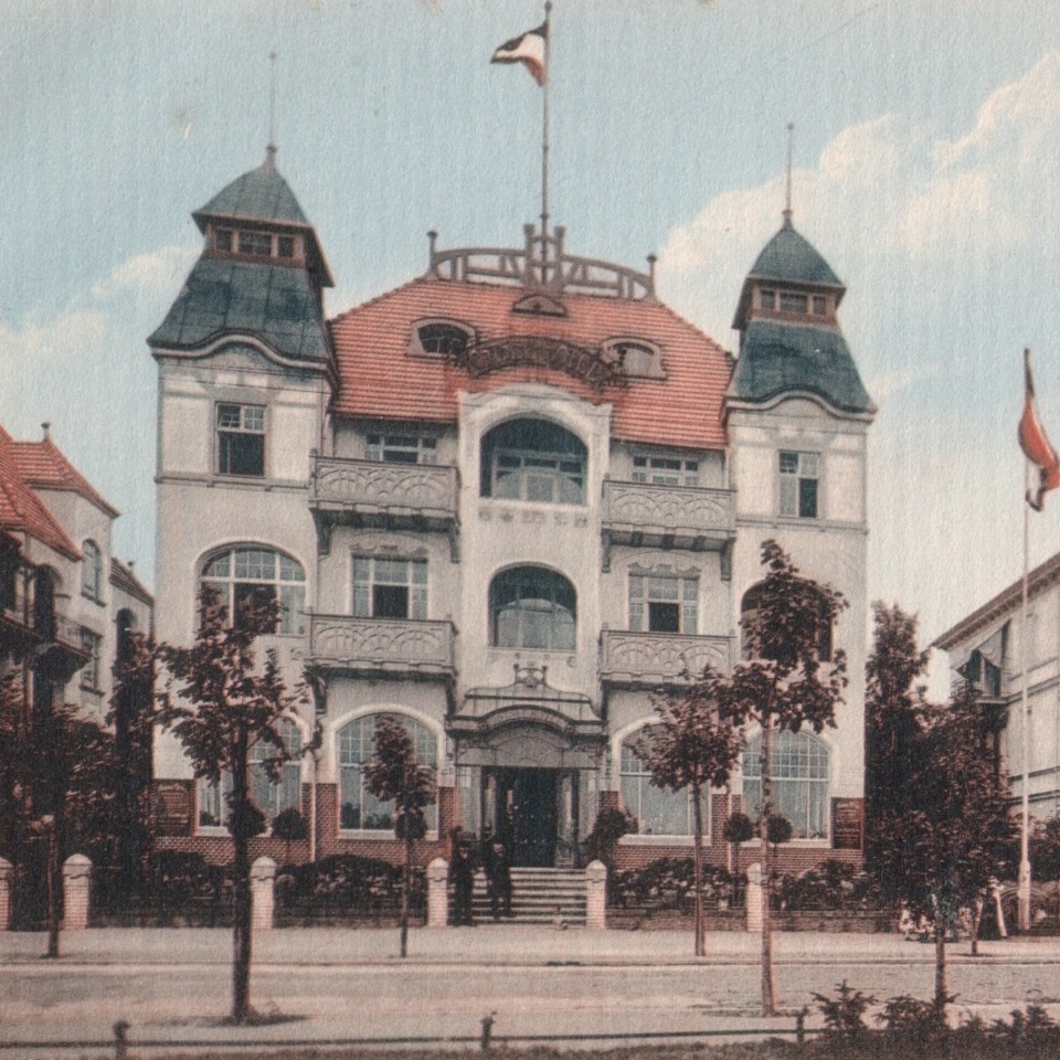 Trzywieżowy hotel o klasycznej architekturze z czerwonym dachem i zielonymi wieżyczkami, otoczony drzewami i ogrodzeniem.