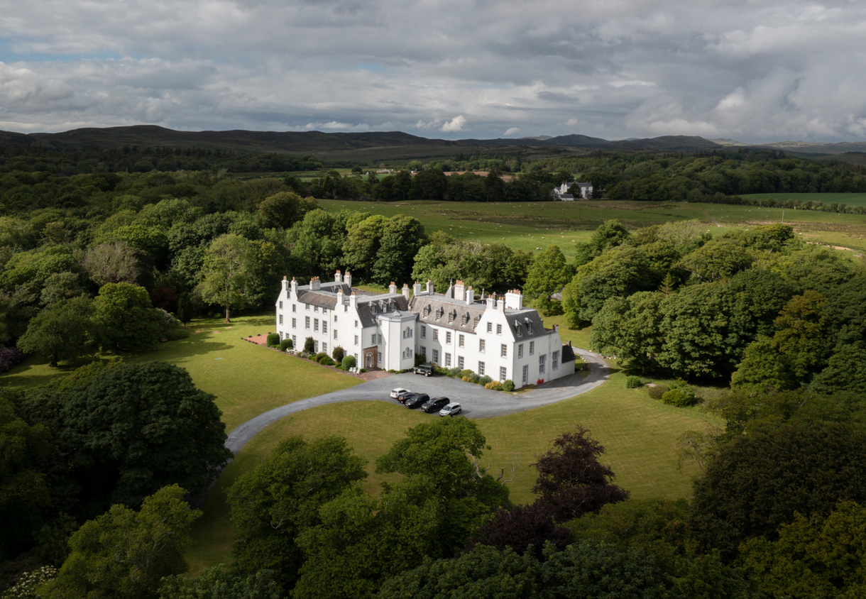 White country manor hotel on expansive lawns, framed by mature woodland, curved gravel drive with parked cars and rolling hills in the distance.