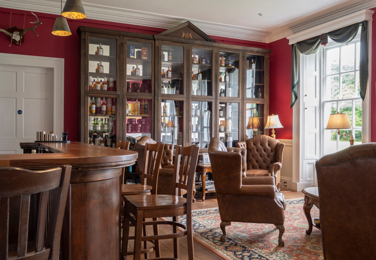 Cozy hotel whisky lounge with dark wood bar, tufted leather armchairs, glass-front liquor cabinet, warm red walls, tall window and table lamps.