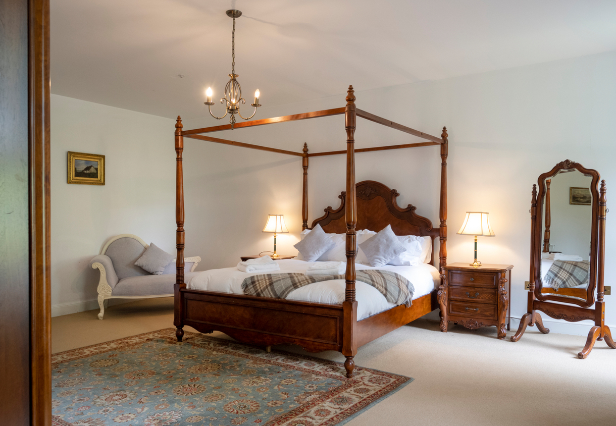 Elegant four-poster wooden bed with white linens, plaid throws, nightstands with lamps, chaise lounge and standing mirror