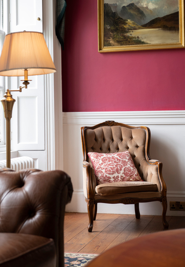Cozy hotel lounge seating with tufted antique armchair and patterned cushion, leather sofa, brass floor lamp, red wall art.