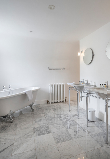 Bright marble bathroom with clawfoot tub by window, twin marble sinks with chrome fixtures, round mirrors and toiletries.