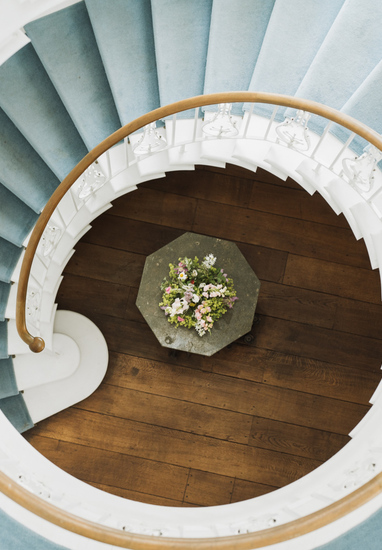 Grand spiral lobby staircase with pale-blue carpet, white balustrade and wood handrail above octagonal table with flowers.