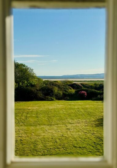 Room window framing manicured lawn and coastal shrubs, with sandy shore and distant sea hills under a clear blue sky