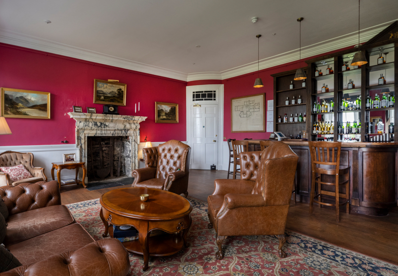 Elegant hotel lounge with tufted leather armchairs, round wooden table, marble fireplace, dark wood bar stocked with spirits.