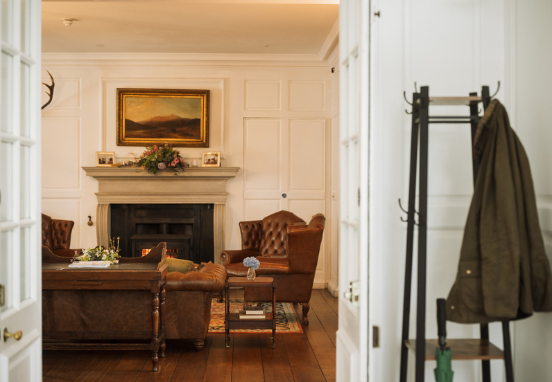 Warm hotel lounge with tufted leather armchairs, lit fireplace, floral mantel, wood floors and coat rack.